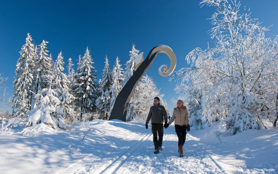 Ein Paar läuft Händchen haltend über den komplett verschneiten WaldSkulpturenWeg in Schanze und im Hintergrund sieht man die Skulptur Krummstab.