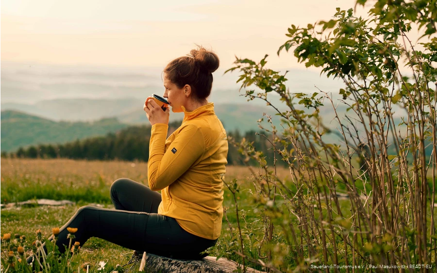 Eine Wanderin genießt ihre Rast bei einer Tasse Kaffee und Blick über die Landschaft.