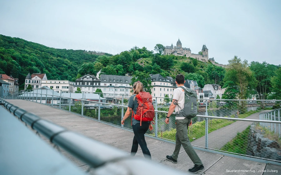 Zwei junge Wanderer überqueren eine Brücke und blicken zur Burg Altena hinauf.
