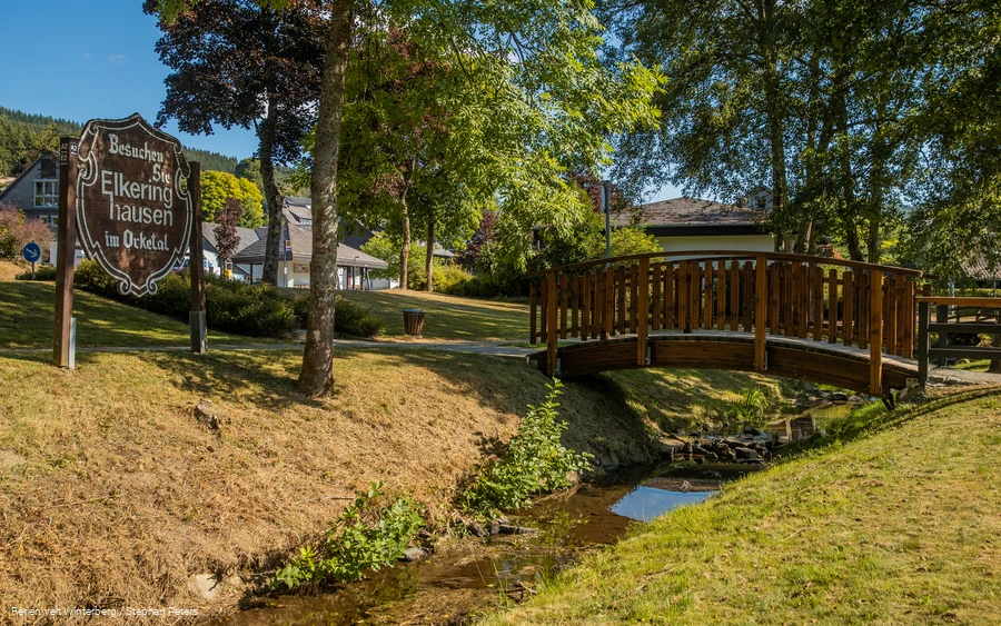 Eine hölzerne Brücke über einem Bach umgeben von Wiese und Bäumen und einem Holzschild mit der Aufschrift Besuchen Sie Elkeringhausen im Orketal.