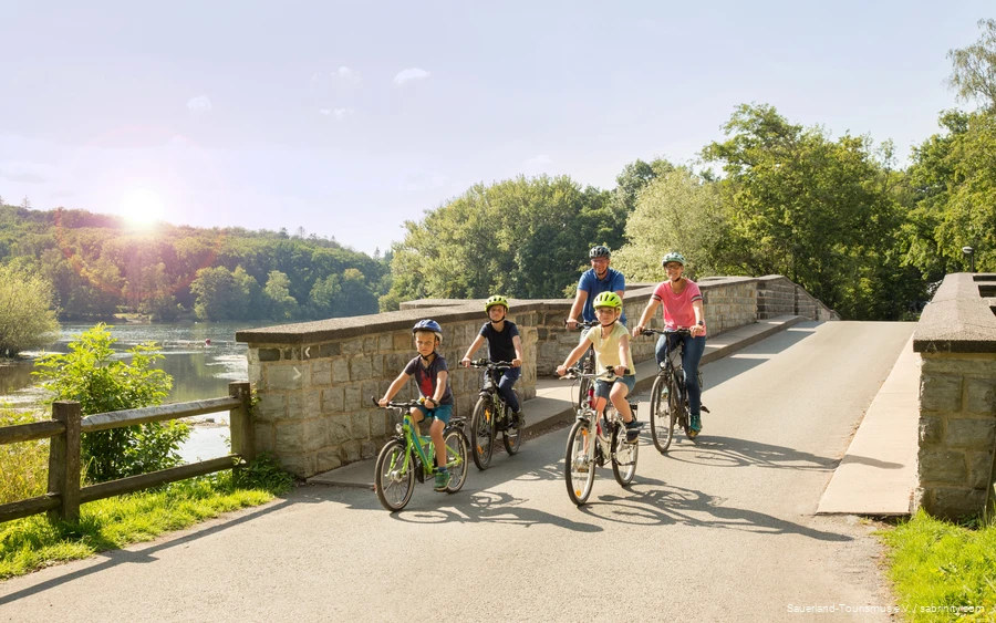 Zwei Eltern und drei Kinder überqueren auf ihren Fahrrädern mithilfe einer Brücke einen Fluss. Die Sonne scheint und die Natur strahlt grün.
