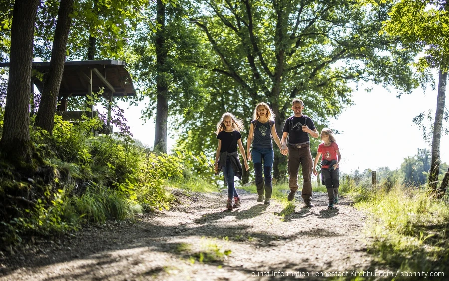 Eine vierköpfige Familie wandert fröhlich und beschwingt einen Wanderweg entlang. Die Eltern halten Händchen, alle lachen.