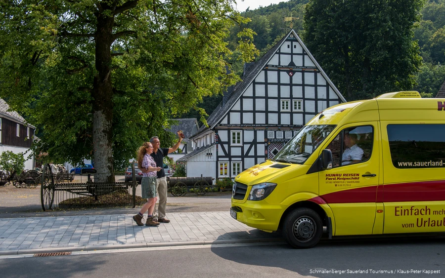 Ein Wanderpärchen winkt einem gelben Mini-Bus, der vor einem Fachwerkhaus geparkt hat.