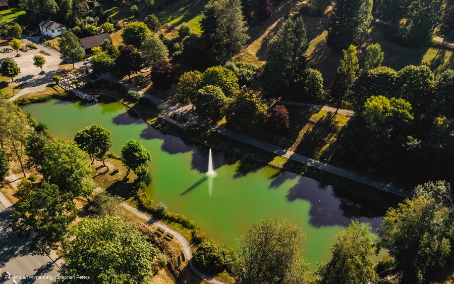 Drohnenaufnahme eines Sees mit einer Fontäne in der Mitte umgeben von Wiesen und Bäumen.