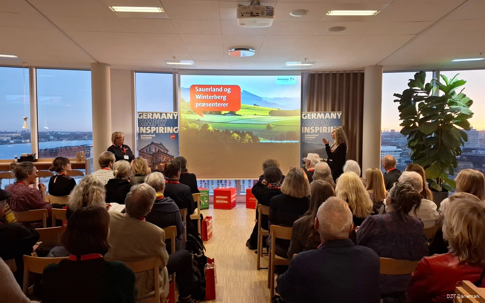 Eine größere Gruppe Menschen schaut auf eine Präsentation. Daneben stehen zwei Frauen, die vortragen.