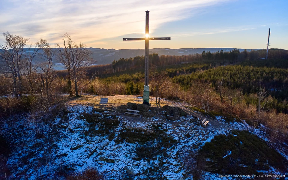 Landschaftsaufnahme von Gipfelkreuz auf dem Olsberg beim Sonnenaufgang. Es liegt eine dünne Schicht Schnee.