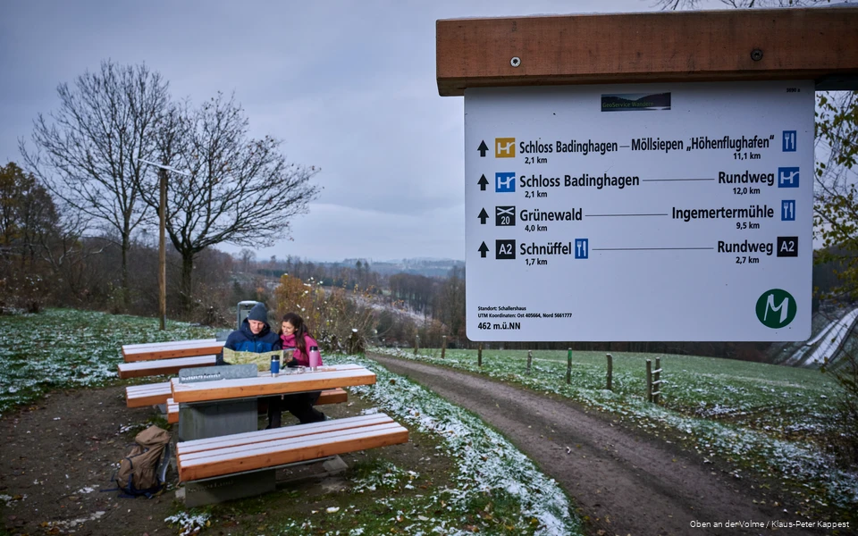 Im Vordergrund ist ein Wegweiser, im Hintergrund sitzt ein Paar auf einer Bank und betrachtet eine Karte. Es liegt eine leichte Schneeschicht.