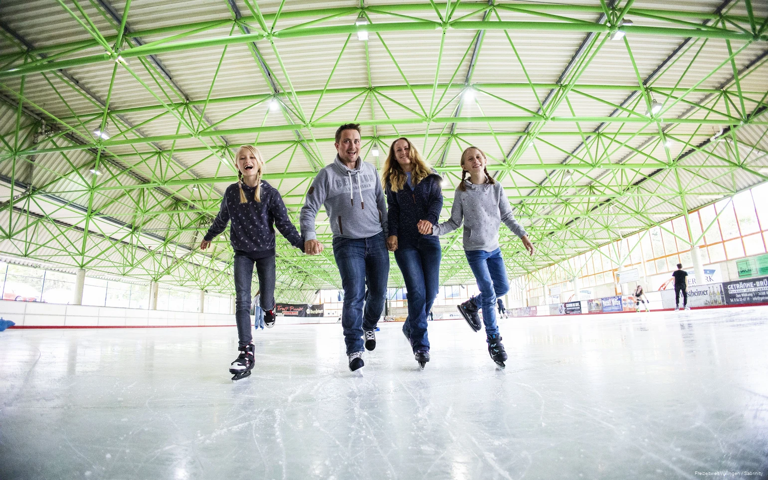 Vater, Mutter und zwei Töchter fahren Hand-in-Hand auf Schlittschuhen.