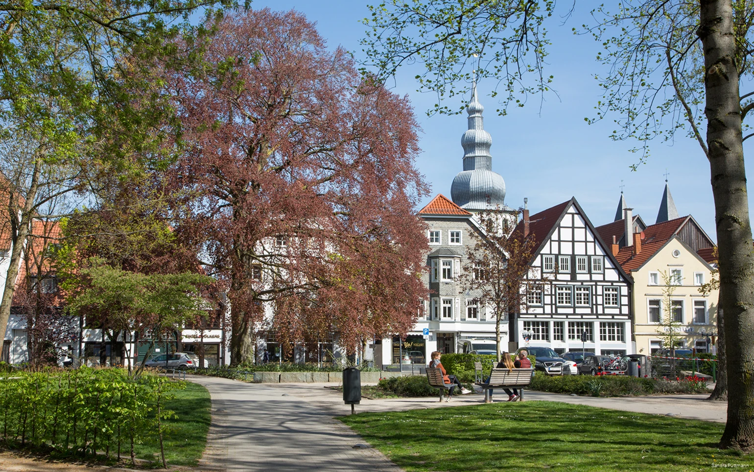 Der Lippstädter Postpark im Frühjahr mit Blick auf die Häuser und Kirchtürme im Hintergrund. Im Vordergrund sitzen drei Menschen auf Bänken.