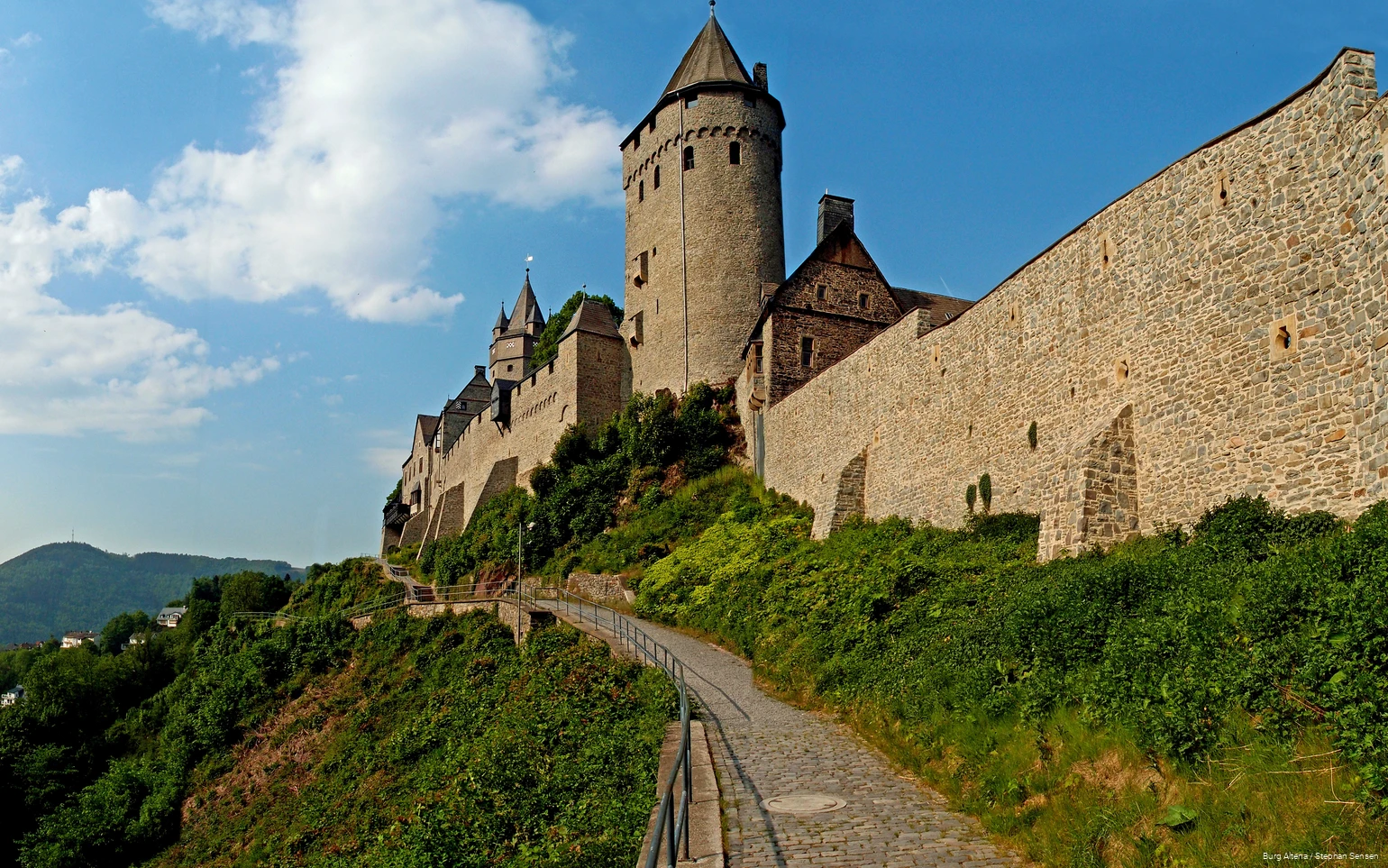 Blick vom Fußweg unterhalb der Burgmauern auf Burg Altena bei blauem Himmel.