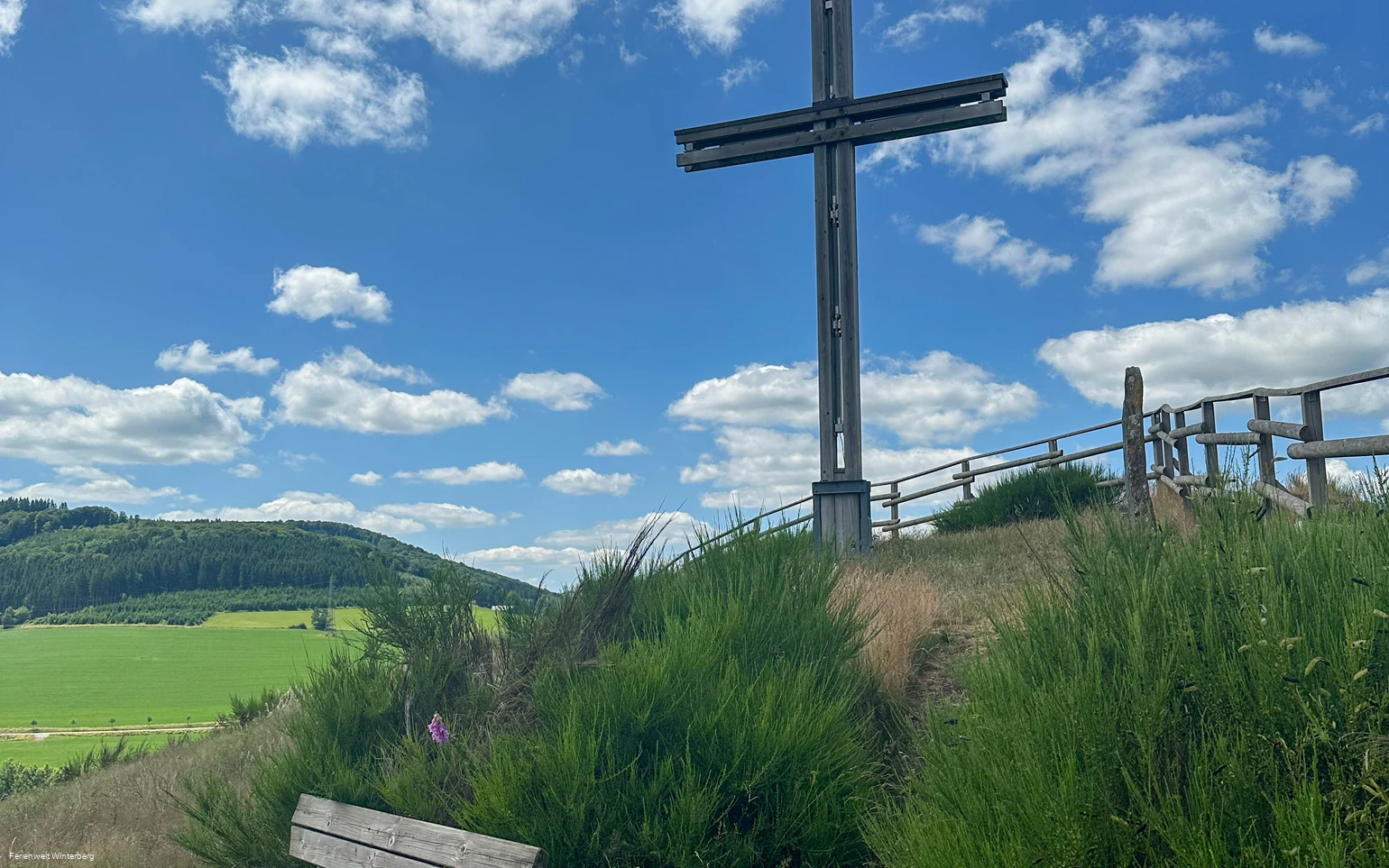 Ein großes Holzkreuz auf einem begrünten Hügel, davor eine Bank.
