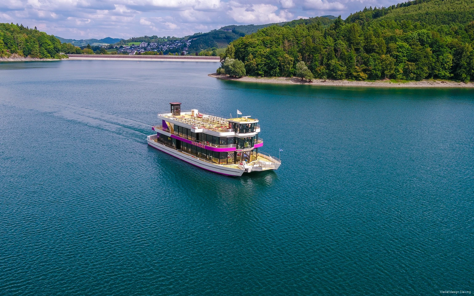 Das Personenschiff EMS Hennesee mit pinken Farbakzenten und einem großen Sonnendeck fährt über den Hennesee. Im Hintergrund ist die Staumauer und ein Teil Meschedes zu sehen.
