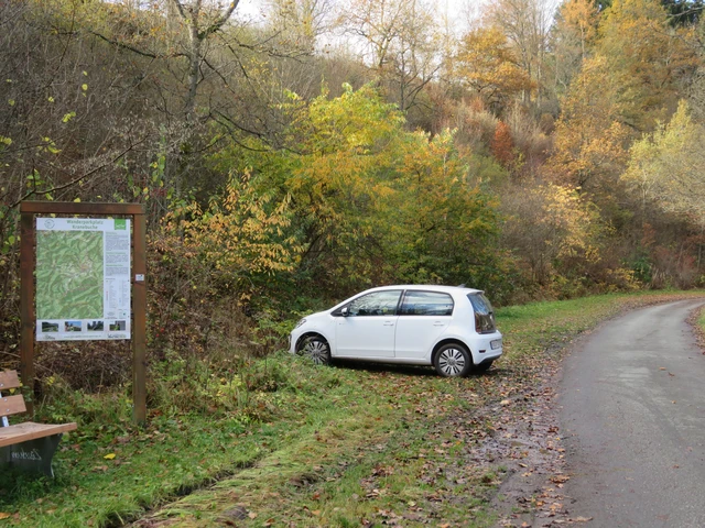 Ein Parkplatz auf dem ein kleines weißes Auto parkt neben einem Waldweg mit einer Infotafel zu Wanderwegen.