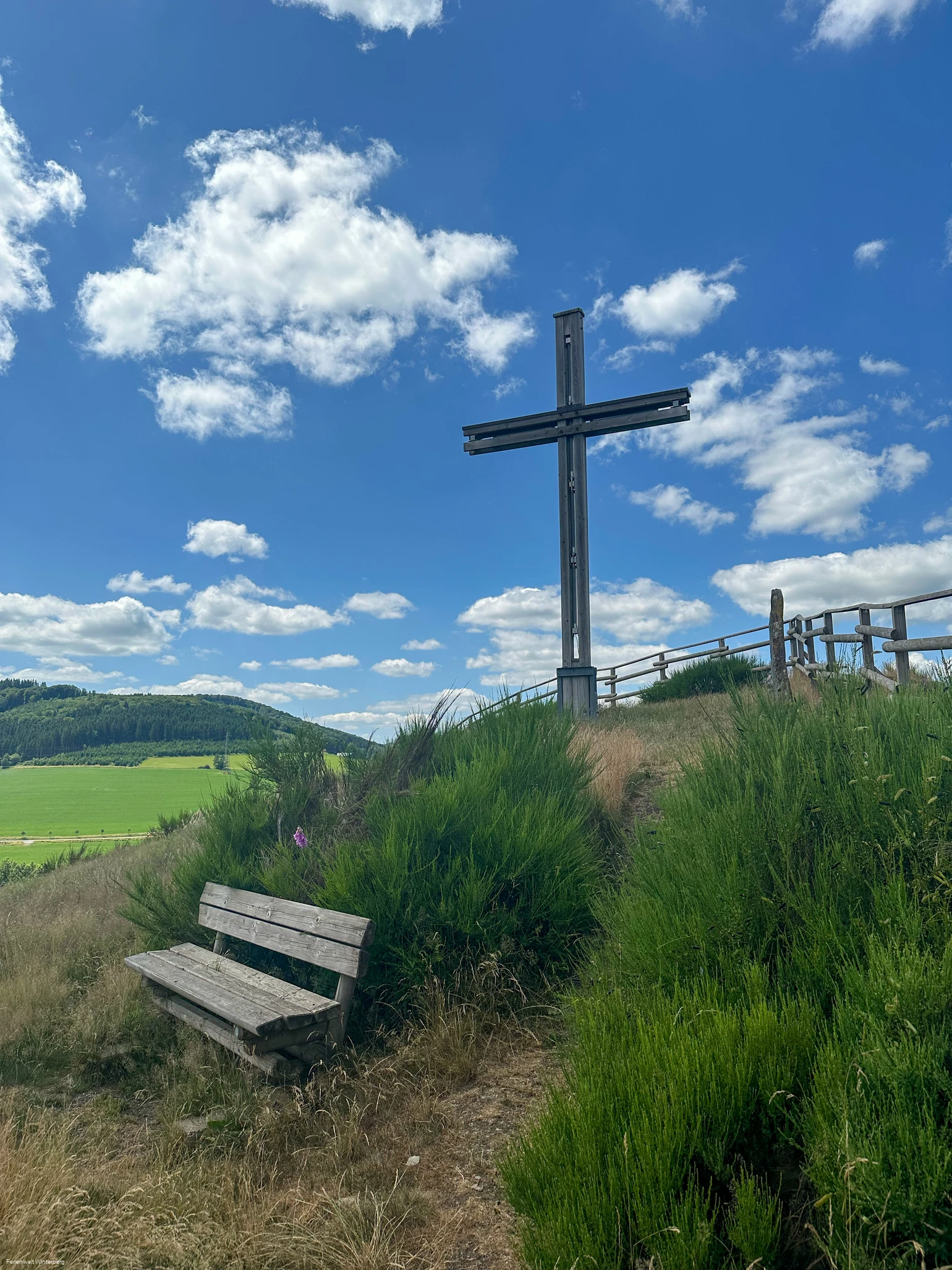 Ein großes Holzkreuz auf einem begrünten Hügel, davor eine Bank.