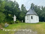Kapelle mit einer Jesusfigur am Kreuz und einer Bank, umgeben von Bäumen.