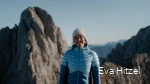 Eva vom Blog Wildwärts grinst mit blauer Daunenjacke und Mütze bekleidet in die Kamera. Im Hinzergrund sind schroffe Berge zu sehen.