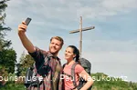 Ein Pärchen in Wanderausrüstung posiert für ein Selfie vor einem Gipfelkreuz.