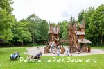 Im Hintergrund spielen Kinder auf einem Spielplatz, der aussieht wie ein Wichteldorf, und im Vordergrund entspannen zwei Väter auf Liegen.
