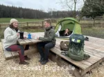 Zwei Wanderer sitzen an einem Tisch an einem Trekkingplatz im Arsnberger Wald. Im Hintergund steht ein Zelt, aus dem eine Frau schaut.