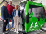 Neben der neu beklebten, grünen Gondel stehen zwei Mitarbeiter vom Sauerland-Tourismus und einer von der Ettelberg-Seilbahn und grinsen.