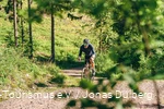 Tätowierter Mann auf einem Gravelbike von vorne. Er fährt einen Schotterweg hoch.