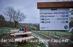In the foreground is a signpost, in the background a couple is sitting on a bench looking at a map. There is a light layer of snow.