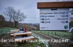 In the foreground is a signpost, in the background a couple is sitting on a bench looking at a map. There is a light layer of snow.