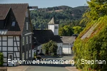 Einige Häuser und eine Kirche an einer Straße mit Bergen und Wäldern im Hintergrund.