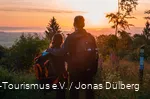 Man and woman stand arm in arm with their backs to the camera and watch a colorful sunset from a vantage point. They are carrying rucksacks and wearing outdoor clothing.