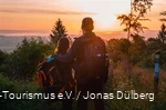 Man and woman stand arm in arm with their backs to the camera and watch a colorful sunset from a vantage point. They are carrying rucksacks and wearing outdoor clothing.