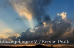 Dunkle Wolken am Abendhimmel mit orangenem Horizont und Sonnenstrahlen, die hinter den Wolken hervorkommen.