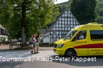 Ein Wanderpärchen winkt einem gelben Mini-Bus, der vor einem Fachwerkhaus geparkt hat.