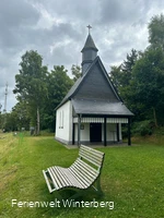 Bank vor einer Kapelle, welche von Bäumen umgeben wird.